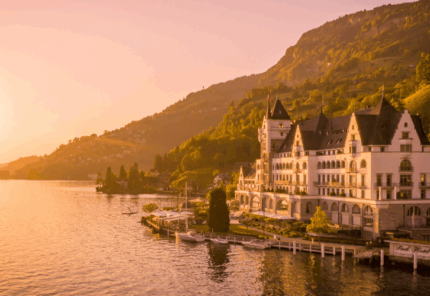A grand lakeside hotel with towers and spires sits at the water’s edge, surrounded by greenery and mountains at sunset, with a warm golden light reflecting on the lake.