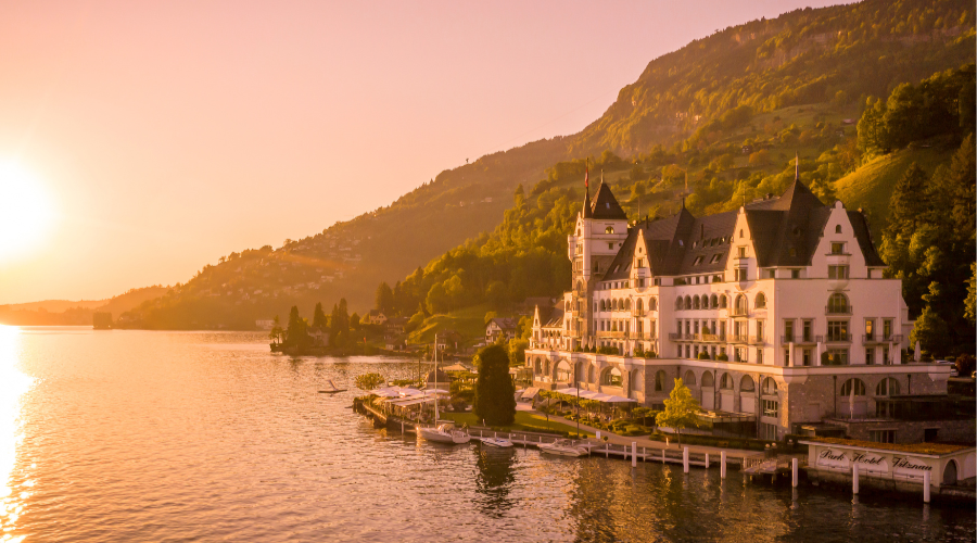 A grand lakeside hotel with towers and spires sits at the water’s edge, surrounded by greenery and mountains at sunset, with a warm golden light reflecting on the lake.