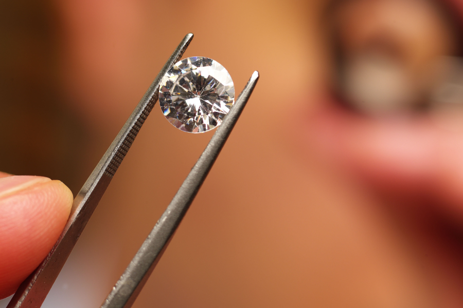 A close-up of tweezers holding a sparkling round-cut diamond, with a blurred background and a persons fingers visible.