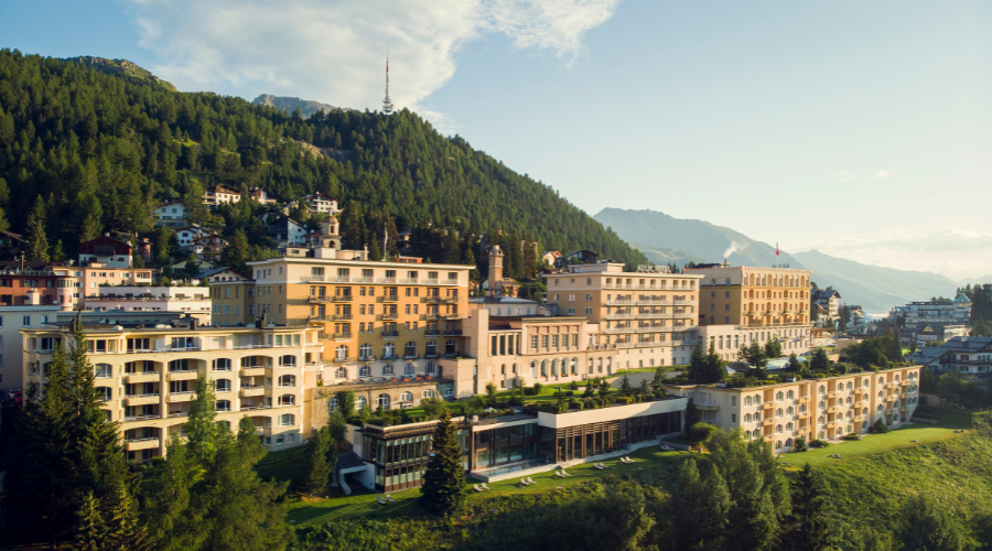 A large, elegant hotel complex with cream-colored buildings sits on a green hillside, surrounded by pine trees and mountains under a clear sky. A communications tower is visible atop the forested hill in the background.
