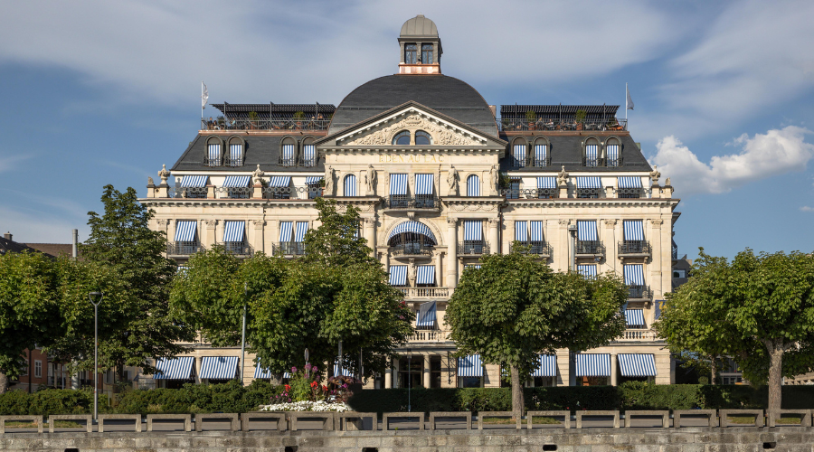 A grand, historic building with ornate architecture, blue-and-white striped window awnings, and a dome on top, stands behind a row of neatly trimmed green trees under a partly cloudy sky.