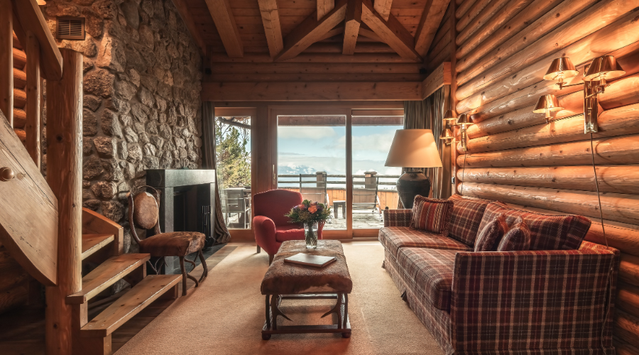 A cozy log cabin living room with a stone fireplace, plaid sofa, and large windows opening to a balcony with a mountain view. Warm lighting and wooden beams create an inviting rustic atmosphere.