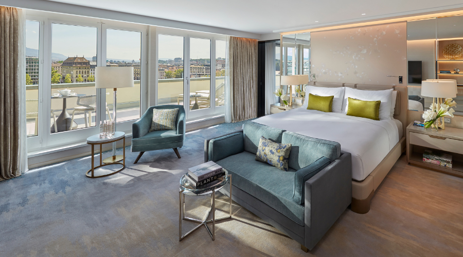 A spacious, modern hotel room with a large bed, two green accent pillows, a loveseat at the foot of the bed, armchair, side tables, and floor-to-ceiling windows overlooking a balcony with a city view.