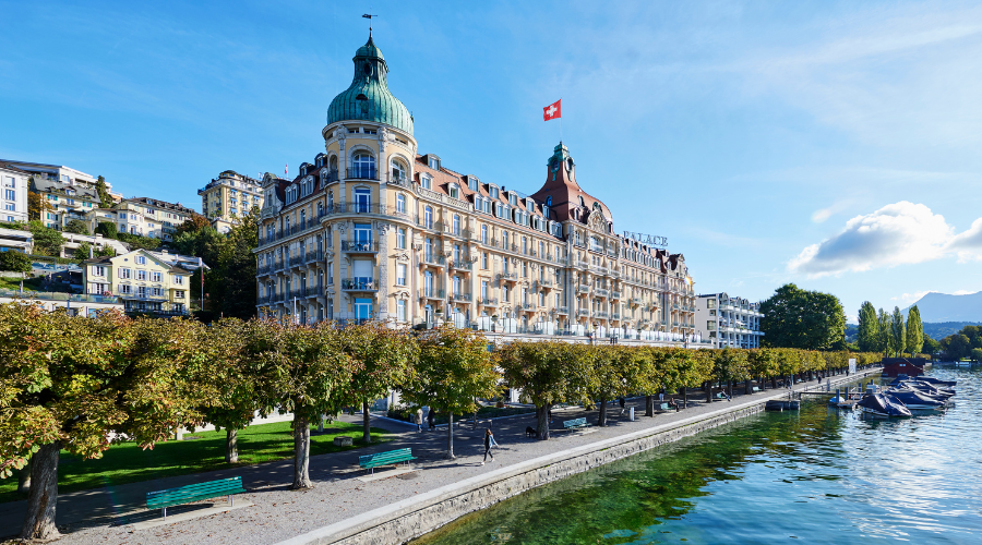A grand historic building with ornate towers and a Swiss flag on the roof stands by a tree-lined promenade next to a clear lake, with benches and boats along the waterfront under a blue sky.