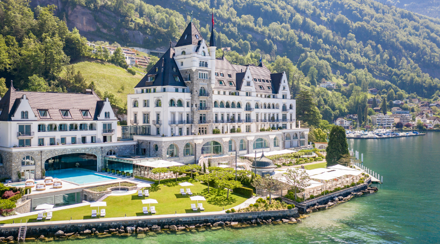 A grand lakeside hotel with turrets and multiple peaked roofs sits beside a pool and manicured lawns, surrounded by green hills and mountains, overlooking clear blue water.