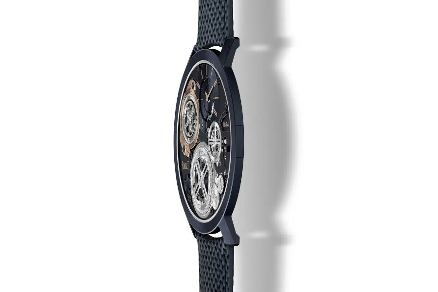 A side view of a luxury wristwatch with a dark textured strap and an intricate, exposed mechanical movement visible through the watch face. The watch casts a soft shadow on a white background.