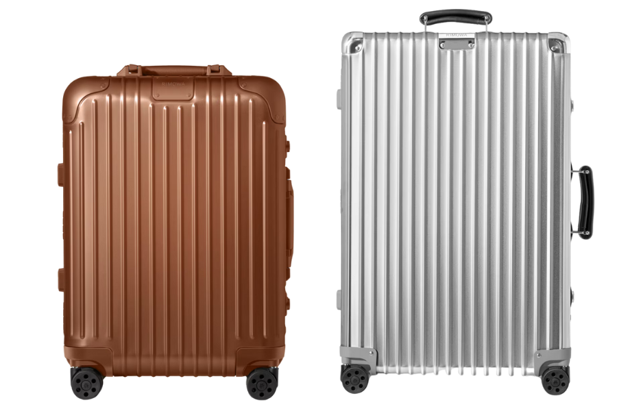 Two hard-shell suitcases with vertical ridges, one in metallic bronze on the left and one in metallic silver on the right, both standing upright with four wheels visible at the base.