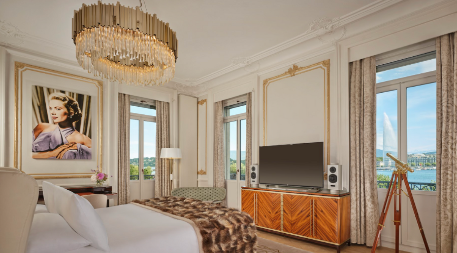 Elegant hotel room with large windows, a chandelier, a bed with a fur throw, a TV on a wooden cabinet, a telescope, and a framed portrait of a woman. Outside, water and greenery are visible.
