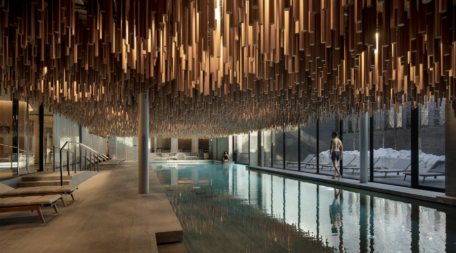 A modern indoor pool with lounge chairs and floor-to-ceiling windows. Warm ceiling lights hang in wooden clusters, creating a striking pattern. Two people relax near the pool, and snow is visible outside.