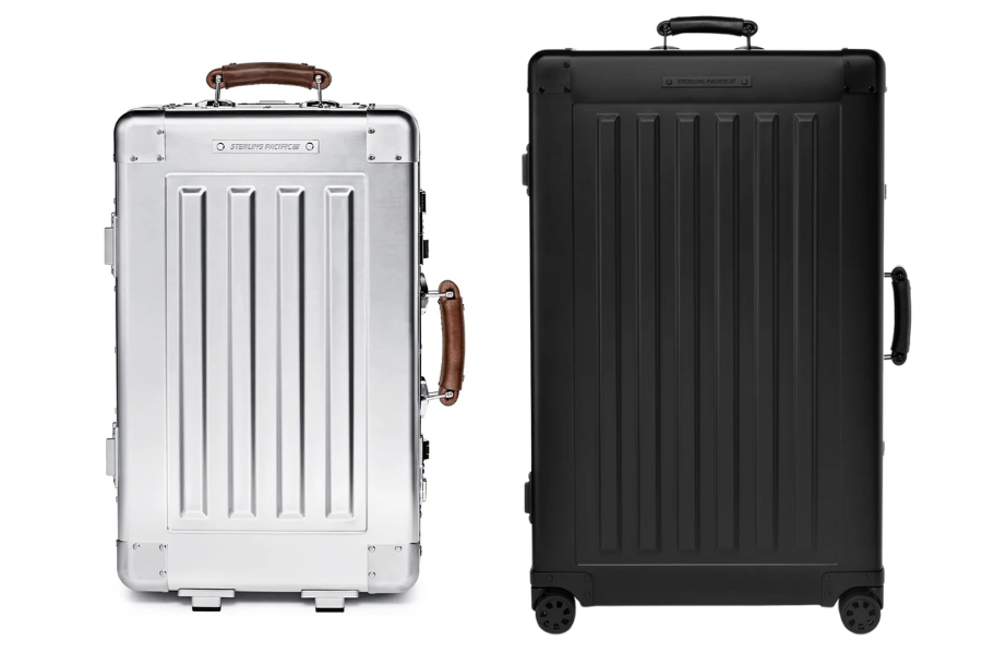 Two upright hard-shell suitcases side by side: one is metallic silver with a brown leather handle, and the other is matte black with matching black handles and wheels. Both have a modern, boxy design.
