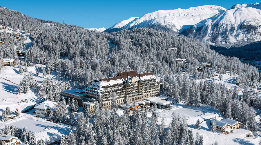 A large historic hotel surrounded by snow-covered trees and buildings, set in a snowy mountain landscape under a clear blue sky.