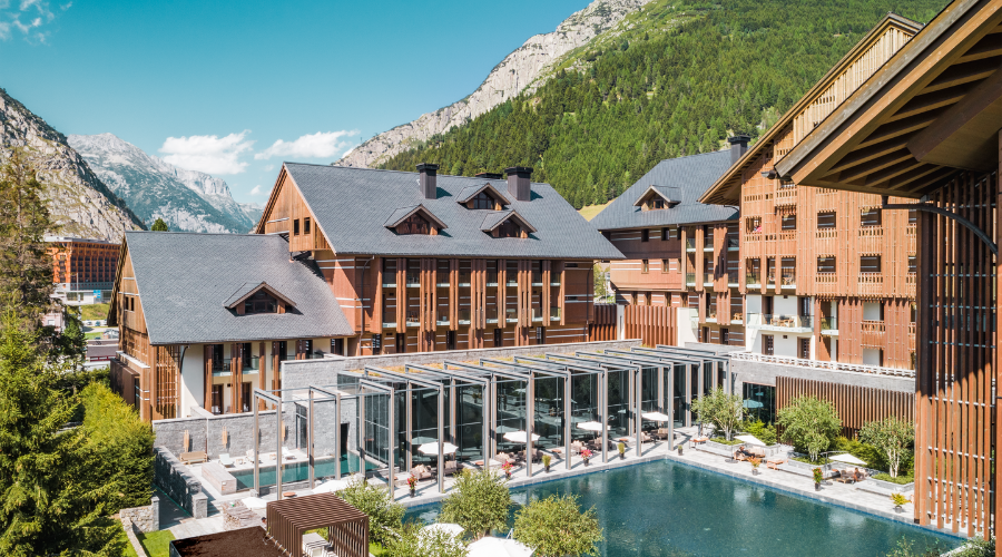 A luxury mountain resort with wooden buildings, outdoor pool, and sun loungers, surrounded by green trees and high, rocky mountains under a clear blue sky.