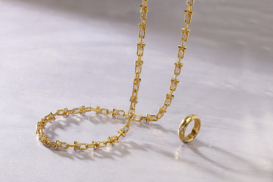 A gold chain necklace with a geometric link design is arranged in a loose spiral next to a thick gold ring on a smooth, light-colored surface.