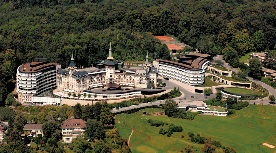 Aerial view of a large, castle-like hotel complex with curved wings, surrounded by trees, tennis courts, and landscaped grounds, located near a forested area.