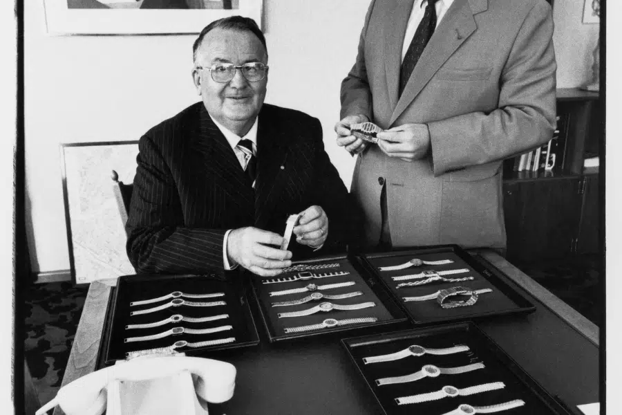 Two men in suits display and examine luxury wristwatches arranged neatly in trays on a table. One man is seated, smiling at the camera, while the other stands beside him holding a watch.