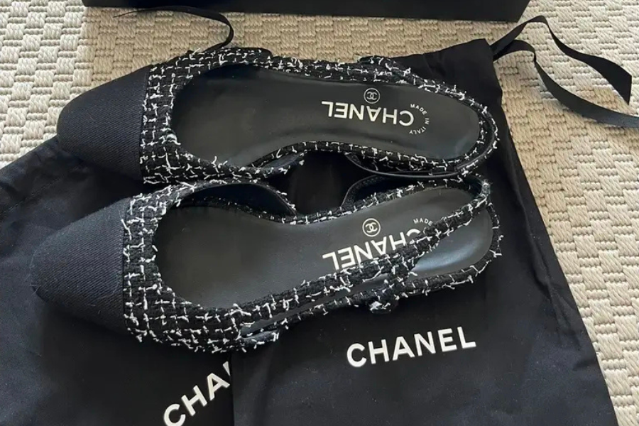 A pair of Chanel slingback shoes with black and white tweed fabric and black toe caps, placed on a black Chanel dust bag on a textured beige surface.