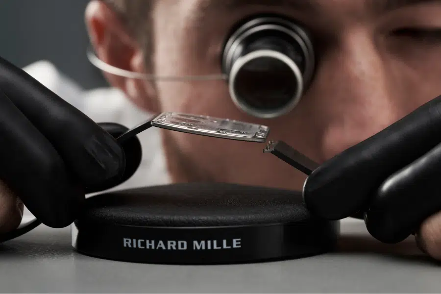 A watchmaker wearing black gloves and a magnifying loupe inspects a watch component on a stand labeled Richard Mille.