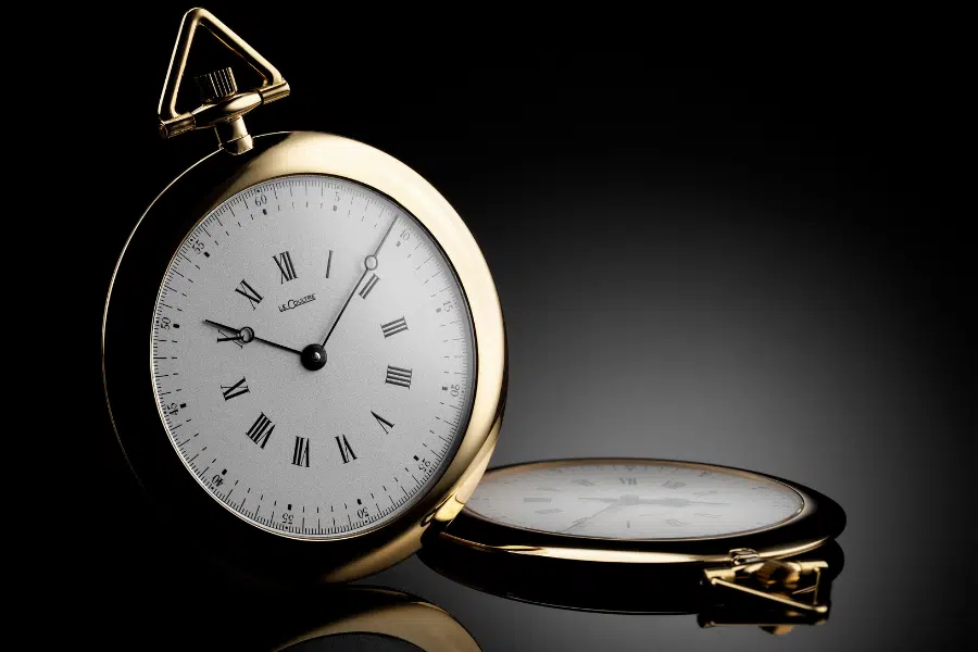 A gold pocket watch with a white face, black Roman numerals, and two black hands stands upright on a reflective surface, with its closed case lying beside it against a dark background.