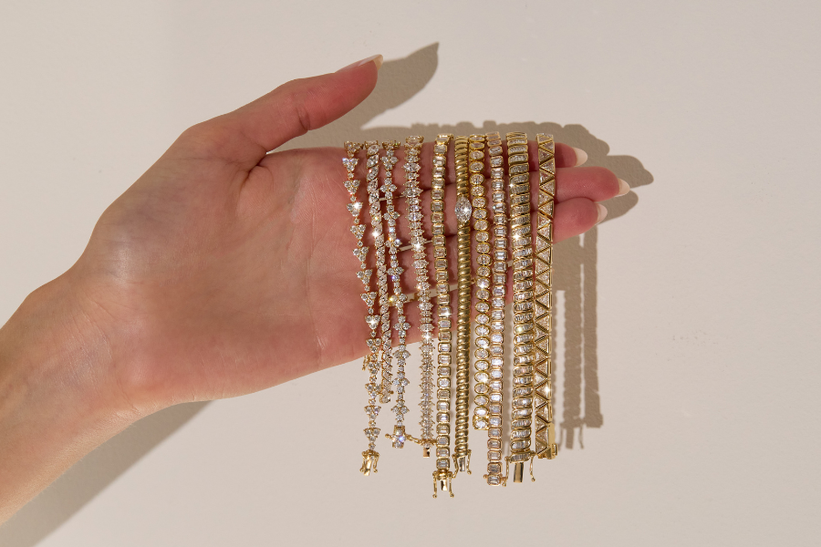 A hand holds several gold and silver bracelets, each adorned with sparkling gemstones and intricate designs, against a neutral background.