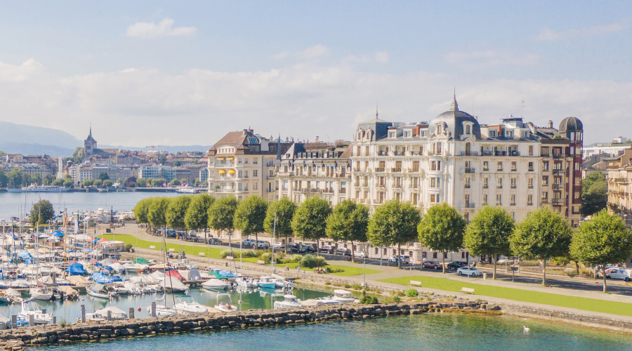 A view of a marina filled with boats, tree-lined paths, and elegant historic buildings along the waterfront under a partly cloudy sky, with a cityscape in the background.