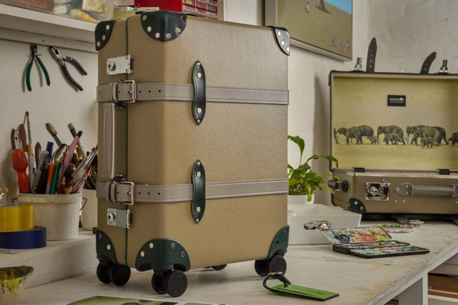 A tan vintage-style suitcase with dark green trim and metal accents stands upright on four wheels in a workspace, with art supplies and another open suitcase in the background.