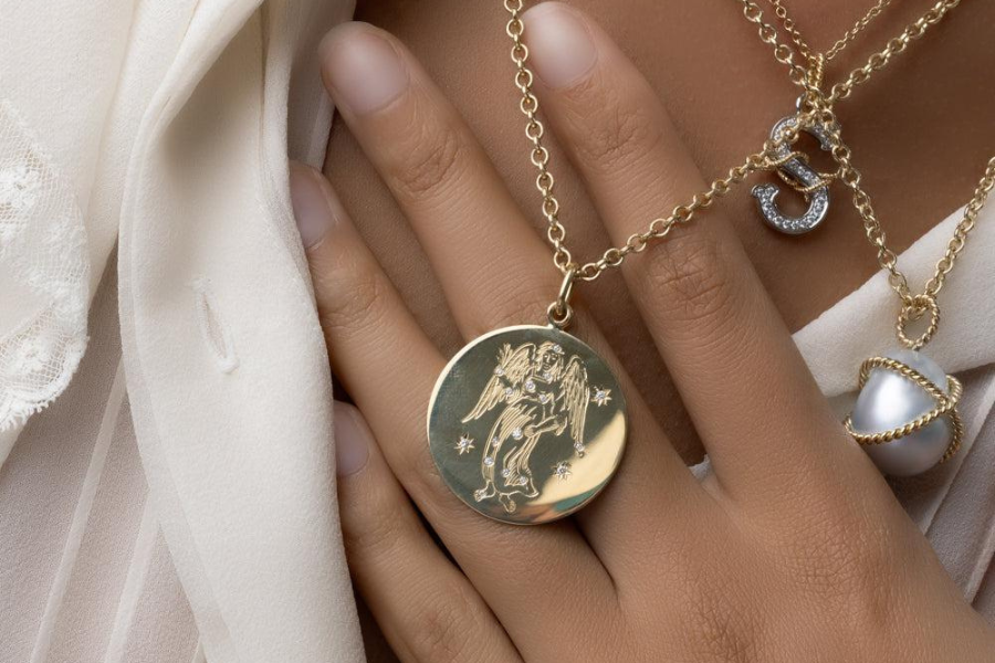 A close-up of a person wearing layered gold necklaces, one featuring a round pendant engraved with an angel, and another with a pearl and a crystal-encrusted letter “S.” The person is touching their chest over a white blouse.