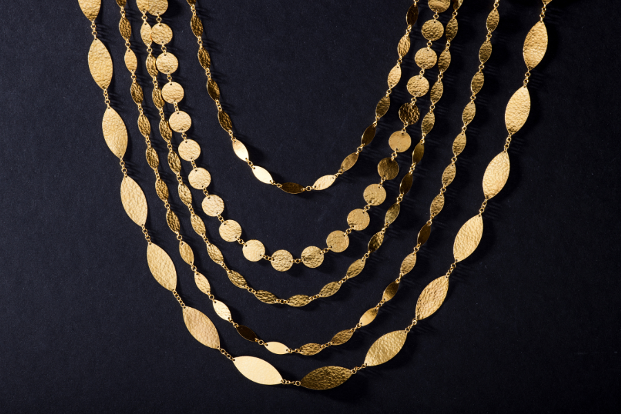 Five gold necklaces with textured, oval and round links are arranged in layers against a black background. The necklaces vary in size, with each lower necklace having larger links than the one above it.