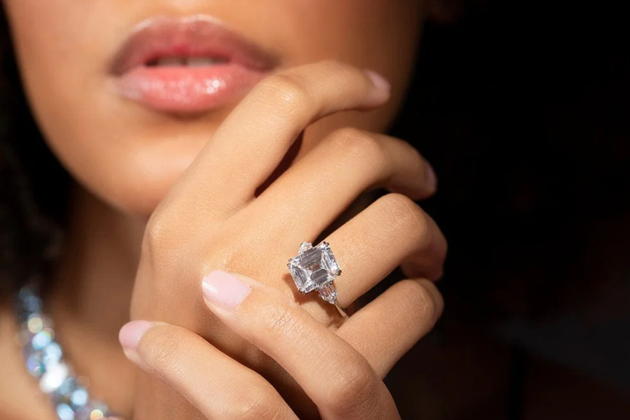 A close-up of a womans hand with manicured nails, showcasing a large square-cut diamond ring on her finger. Her lips and part of a sparkly necklace are visible in the background.