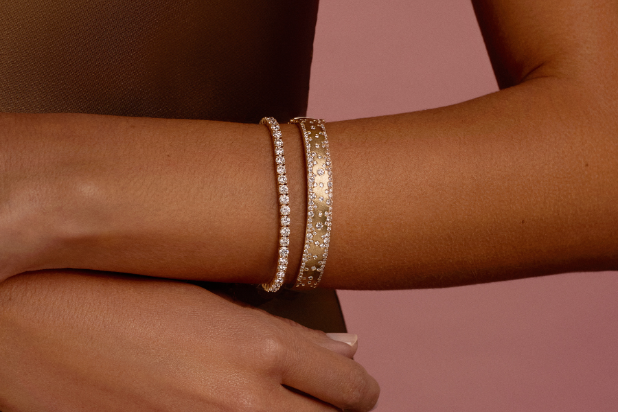 A close-up of a person’s forearm wearing two elegant gold bracelets, one thin with diamonds and one wider with engraved patterns and small stones, against a soft pink background.