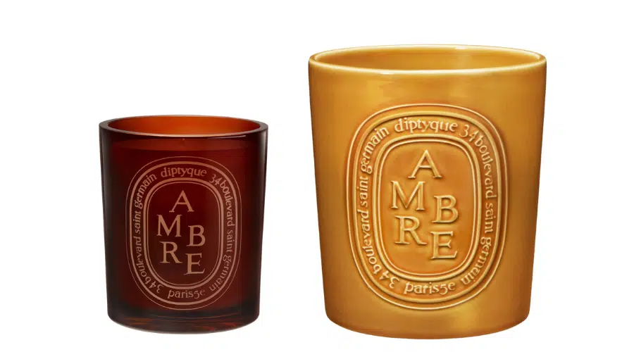 Two Diptyque Ambre candles are shown: one in a small, dark amber glass holder and the other in a larger, matte yellow ceramic holder. Both feature an embossed oval label with the word AMBRE.
