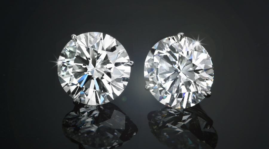 Two round, brilliant-cut diamonds are set in prongs against a dark, reflective background. Both diamonds sparkle, showcasing their clarity and facets. Their reflections are visible below each stone.
