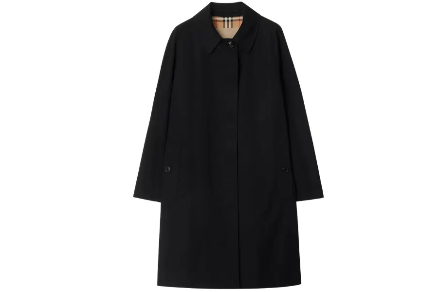 A long black trench coat with a classic collar, button closure, and side pockets. The interior lining features a beige plaid pattern. The coat is displayed against a white background.
