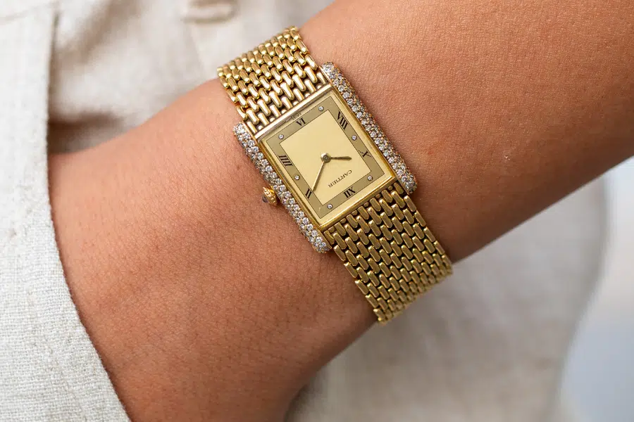 A close-up of a gold rectangular wristwatch with a mesh band and diamond-studded bezel, worn on a persons wrist. The watch face has simple black hour markers and hands, with the brand name Seiko visible.