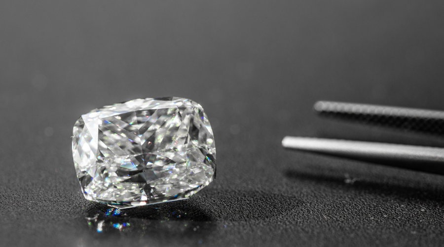 A clear, cushion-cut diamond rests on a dark, textured surface beside a pair of metal tweezers. The diamond sparkles, reflecting light and revealing its intricate facets.