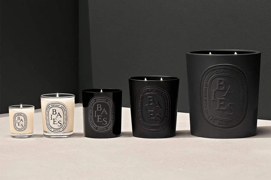Five Diptyque Baies candles in varying sizes, with three black jars and two white jars, are arranged in a row on a light surface against a dark background. All candles are lit.