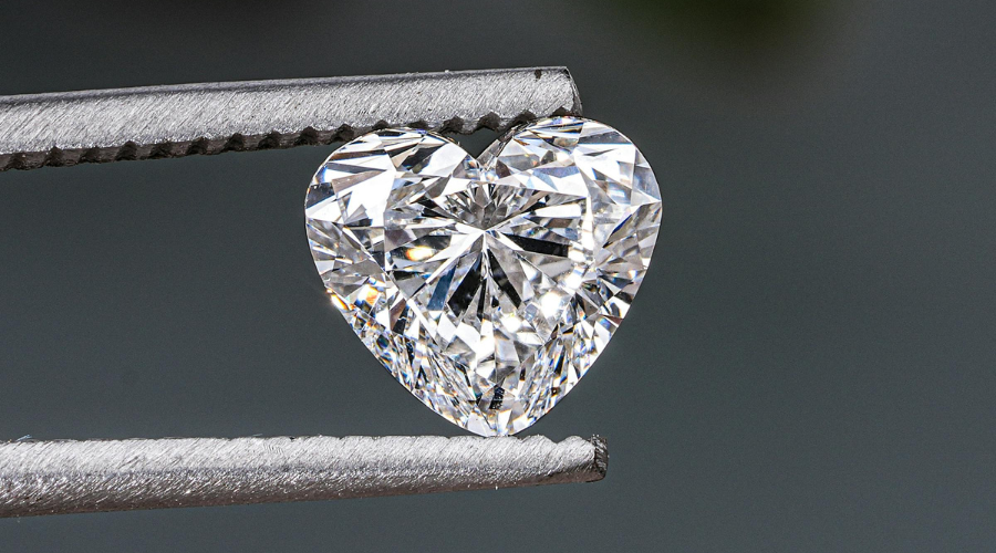 A pair of tweezers holds a sparkling, heart-shaped diamond against a blurred gray background, showcasing the stones clear facets and brilliance.