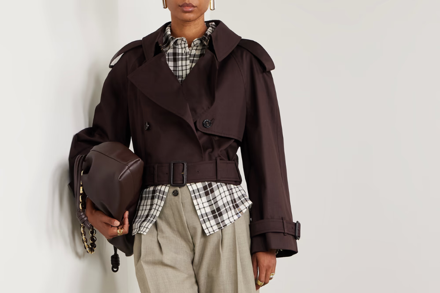 A person wearing a dark cropped trench coat over a black-and-white plaid shirt, beige trousers, and holding a brown bag with a gold chain strap, standing against a plain light background.