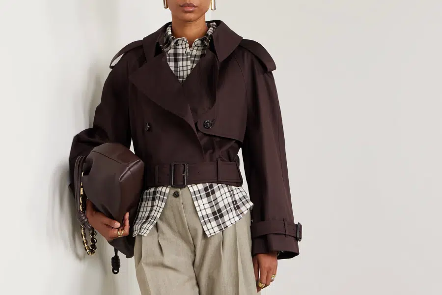 A person wearing a dark cropped trench coat over a black-and-white plaid shirt, beige trousers, and holding a brown bag with a gold chain strap, standing against a plain light background.