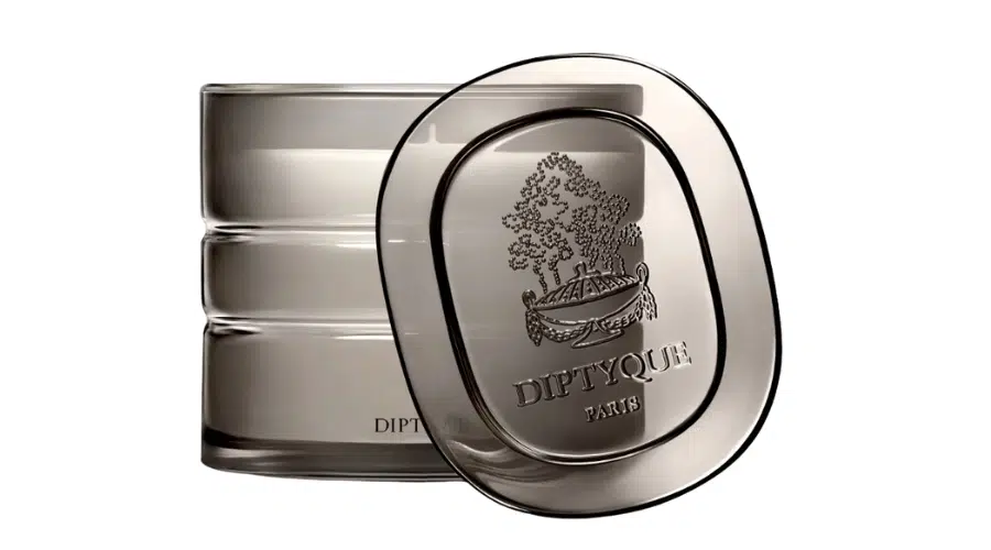 A translucent gray Diptyque Paris candle holder with a matching lid featuring the Diptyque logo and an illustrated scene. The lid is resting tilted against the side of the holder.