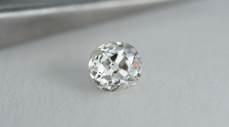 Close-up of a round, faceted, transparent gemstone with a brilliant cut, resting on a white surface with a blurred metallic object in the background.