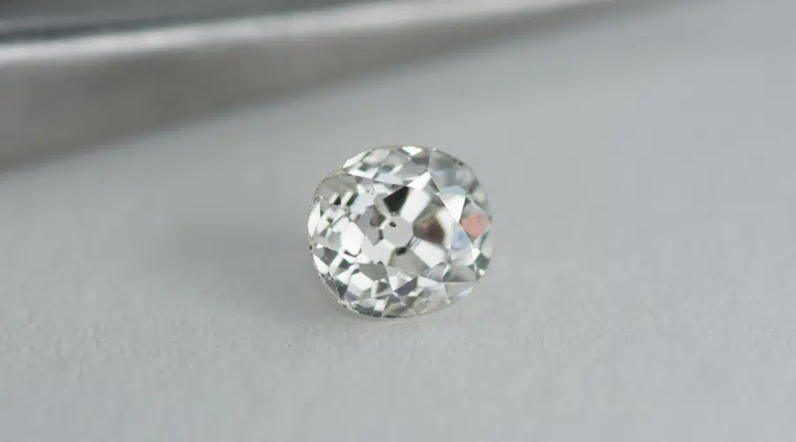 Close-up of a round, faceted, transparent gemstone with a brilliant cut, resting on a white surface with a blurred metallic object in the background.
