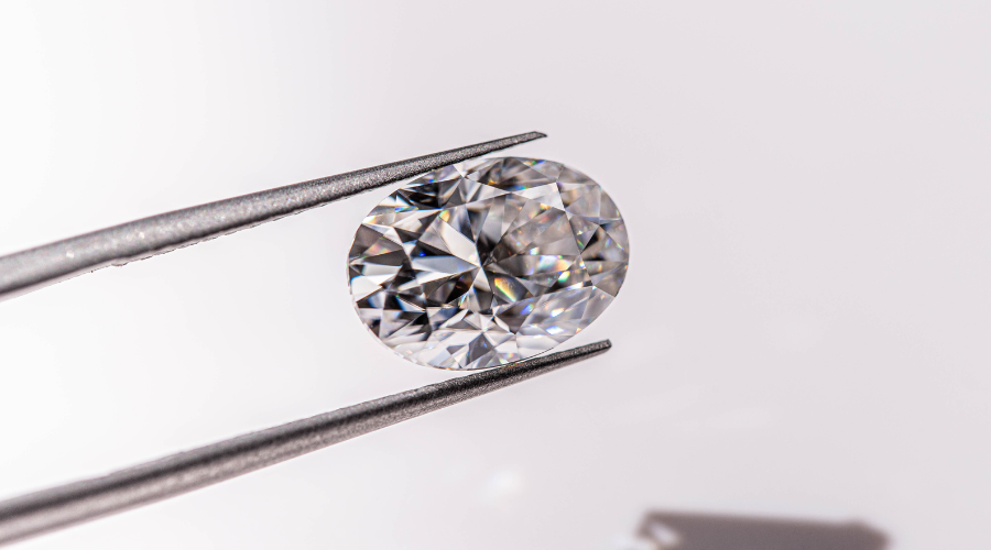 A pair of tweezers holding a sparkling, oval-cut diamond against a white background, with light reflecting off the diamond’s facets.