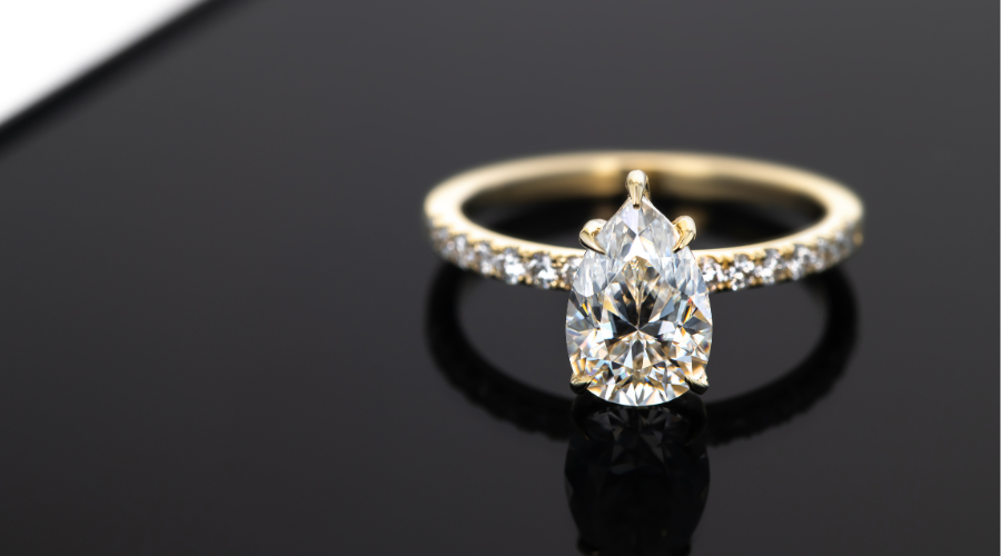 A gold ring with a large pear-shaped diamond center stone and a band adorned with smaller diamonds rests on a glossy black surface.