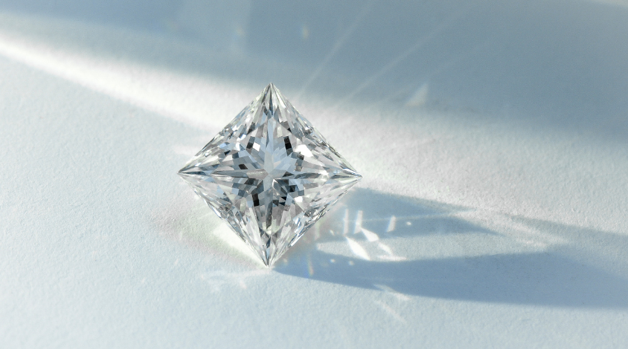 A clear, sparkling diamond with a square pyramid shape sits on a light surface, casting intricate reflections and shadows in the soft light.