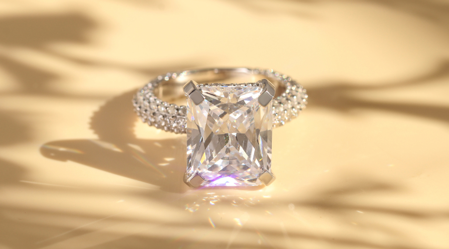 A radiant-cut diamond ring with a sparkling, cushion-shaped center stone and a pavé diamond band, displayed on a light beige surface with soft shadow patterns.