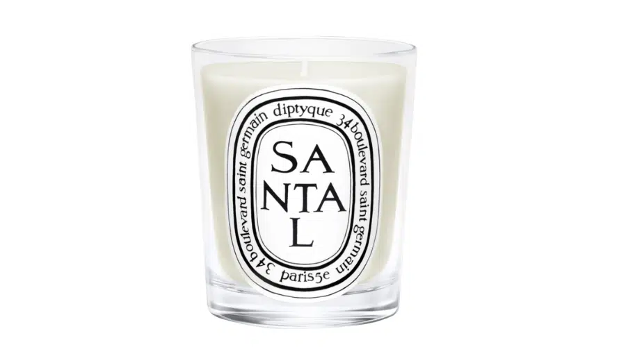 A white candle in a clear glass holder with an oval Diptyque label reading SANTAL and the brand’s name in black text around the border.