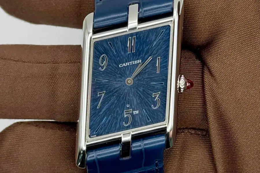 A rectangular Cartier wristwatch with a blue textured face, silver hands, and numerals 3, 5, 7, 9, and 11. The watch has a blue leather strap and is held by a gloved hand.
