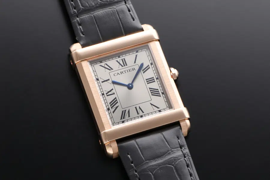 A luxury rectangular wristwatch with a gold case, white dial, black Roman numerals, blue hands, and a black leather strap, displayed on a dark surface.