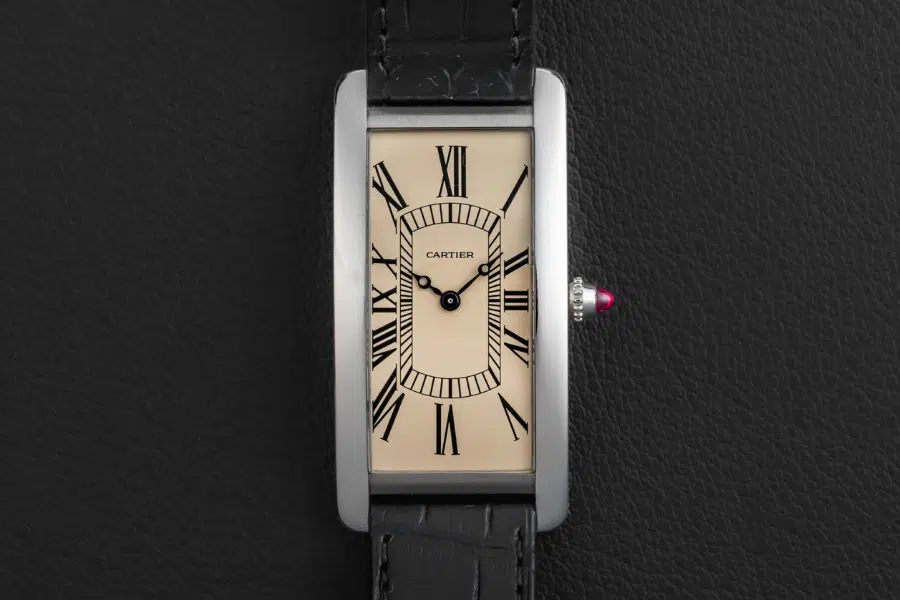 A rectangular Cartier wristwatch with a cream dial, black Roman numerals, blue steel hands, and a black leather strap displayed on a textured black surface.