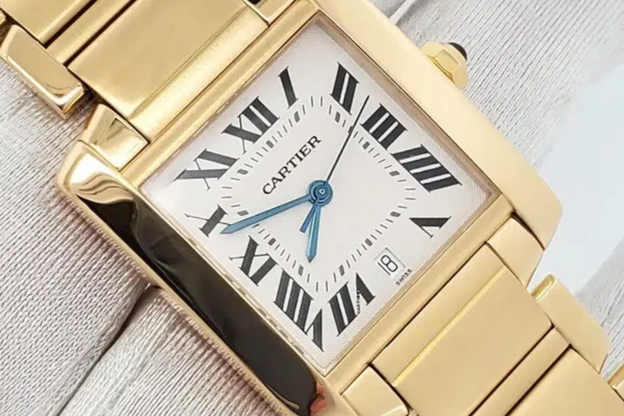 Close-up of a gold Cartier wristwatch with a square face, Roman numerals, blue hands, and a date window at the 3 oclock position, resting on a light-colored fabric background.